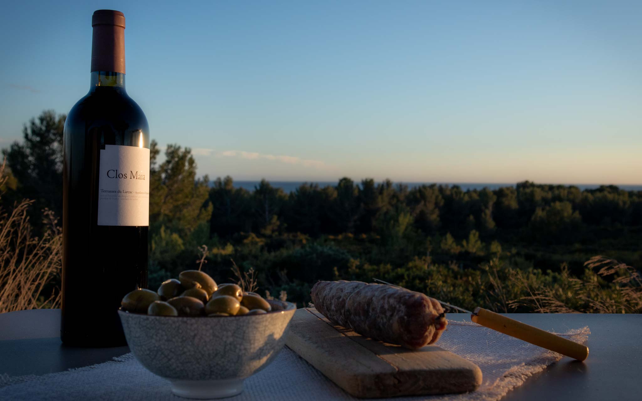 Bouteille de Clos Maïa Rouge un vin des Terrasses du Larzac avec des olives et un saucisson